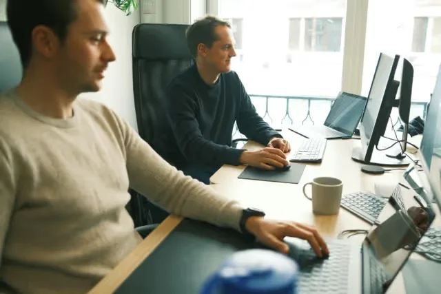 Two men working at desks with computers in a bright office space.