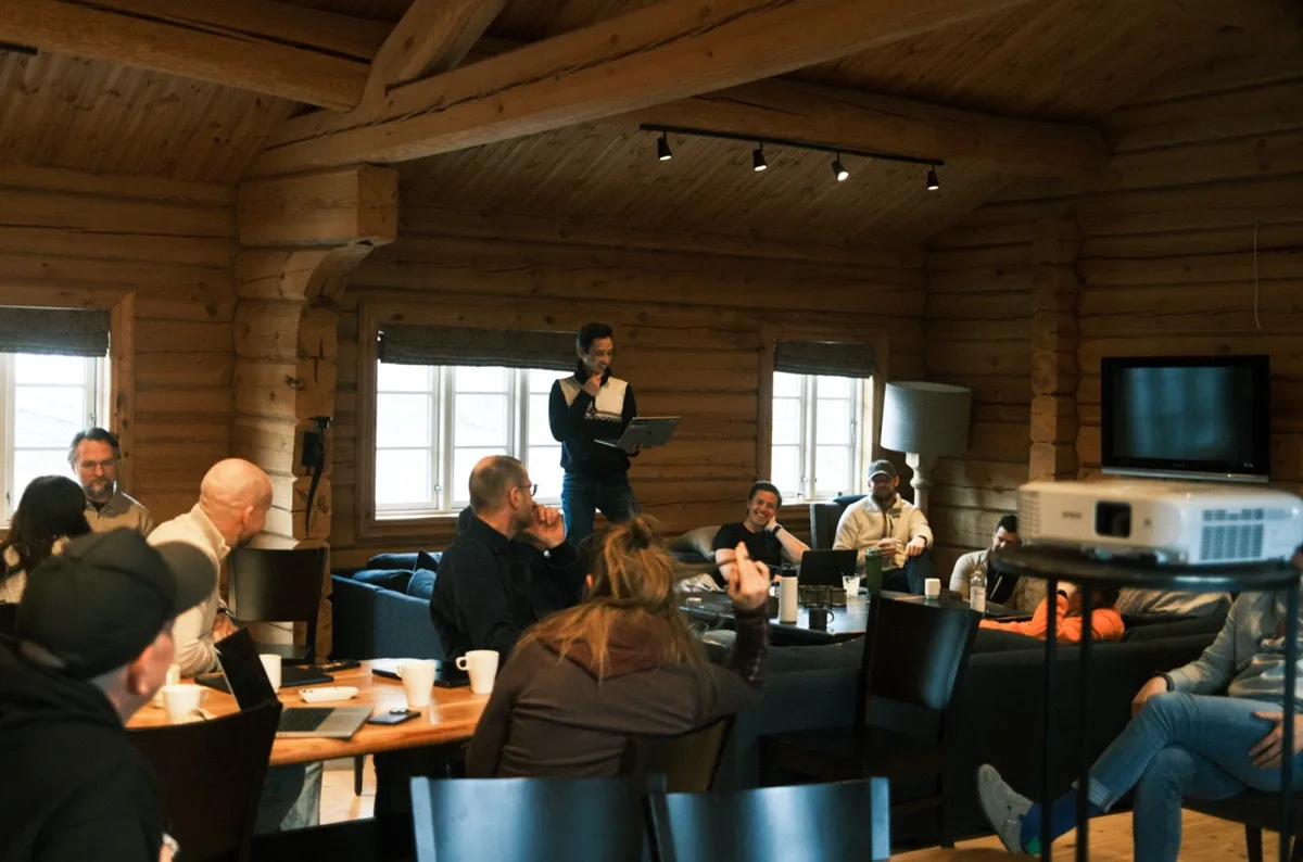 Group of people sitting and listening to a man standing and speaking in a cozy wooden room with large windows and a projector.