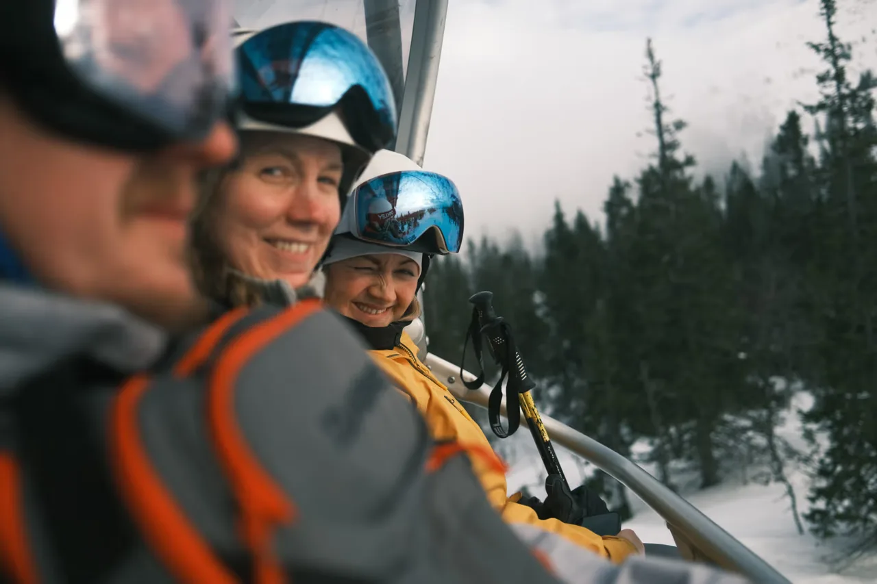 Three skiers wearing helmets and goggles sitting on a ski lift above a snowy forest.