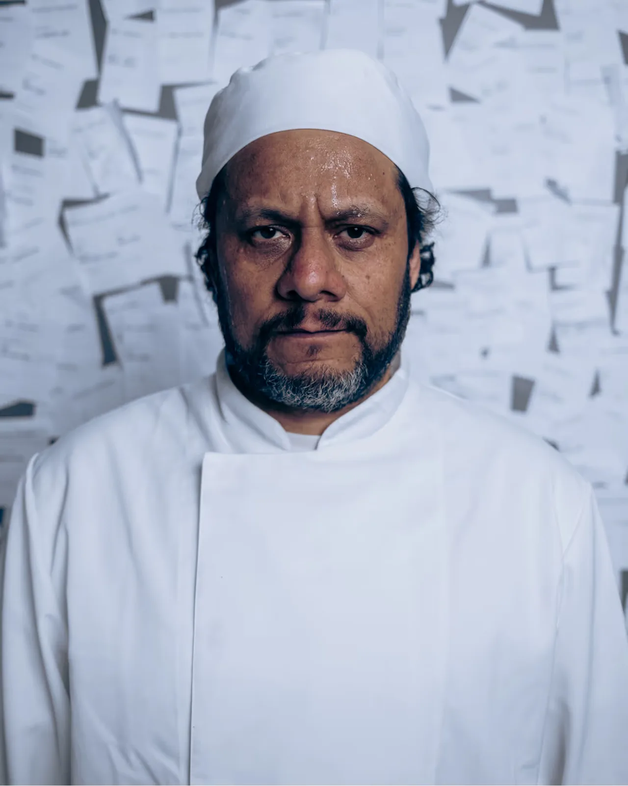 Chef in white uniform and hat stands against a wall covered with pinned papers.