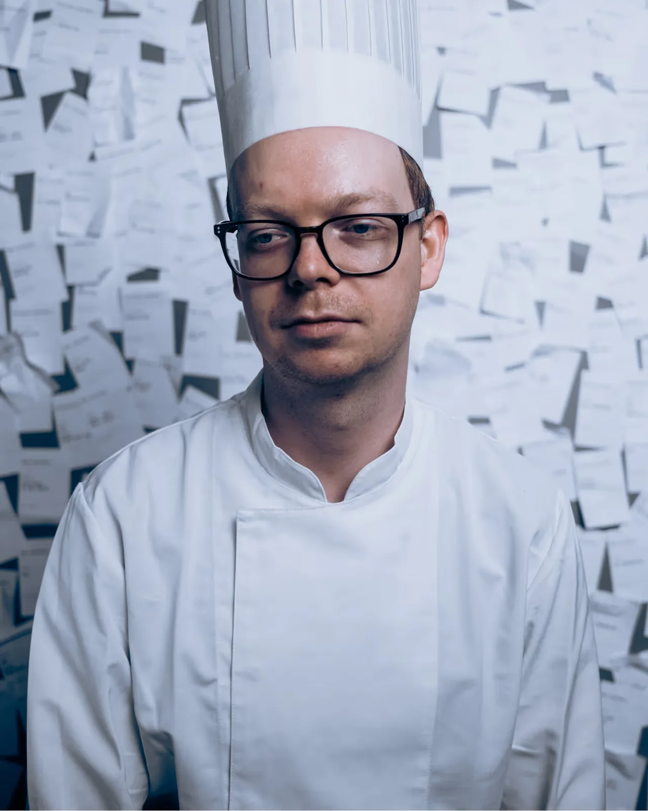 Man in white chef uniform and tall hat standing in front of a wall covered with papers.