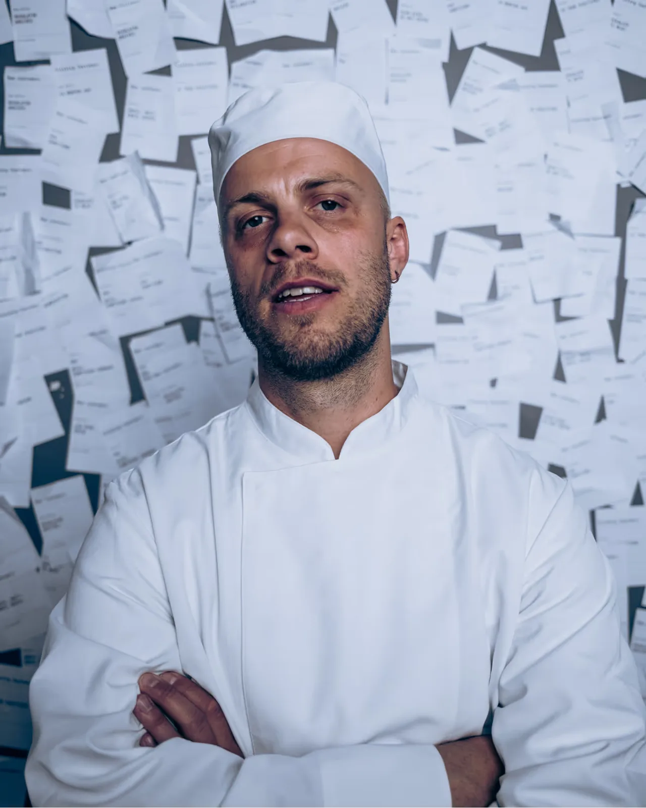Male chef in white uniform and hat stands with arms crossed against wall covered in papers.