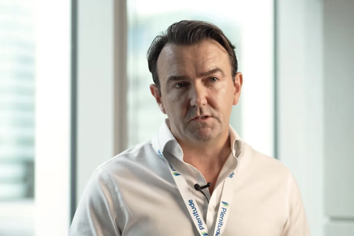 Man in white shirt wearing a lanyard with ‘Plenitude’ logo, speaking indoors with blurred windows in the background.