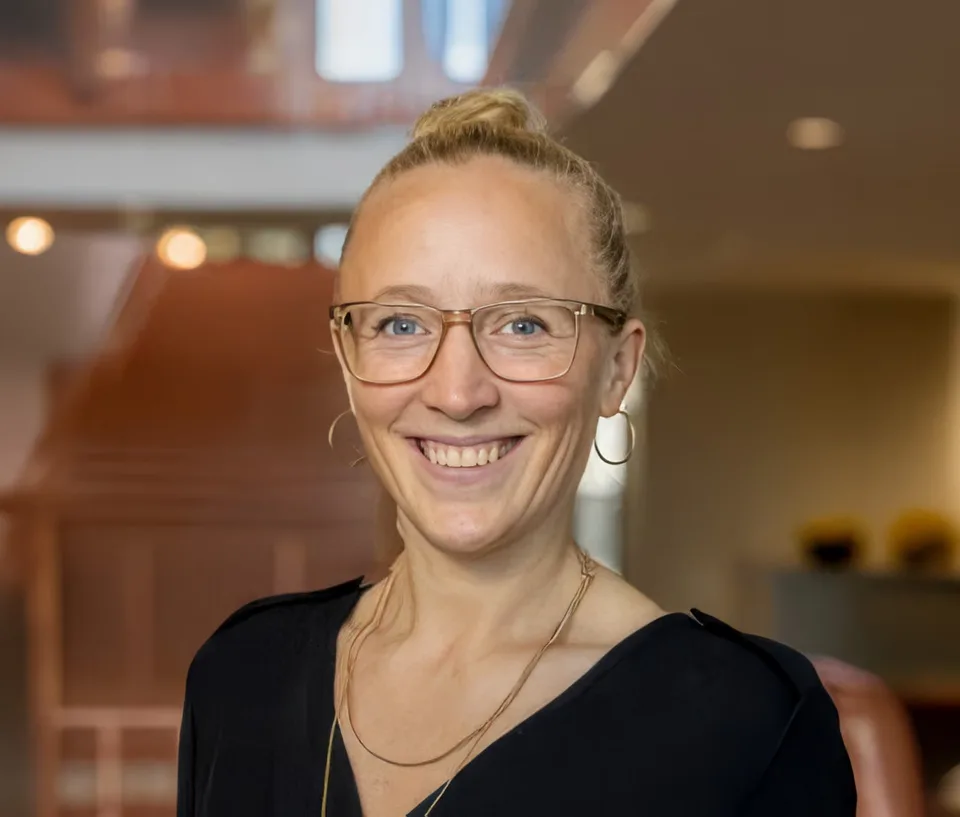 Smiling woman with glasses and blonde hair in a bun, wearing hoop earrings and a black top indoors.