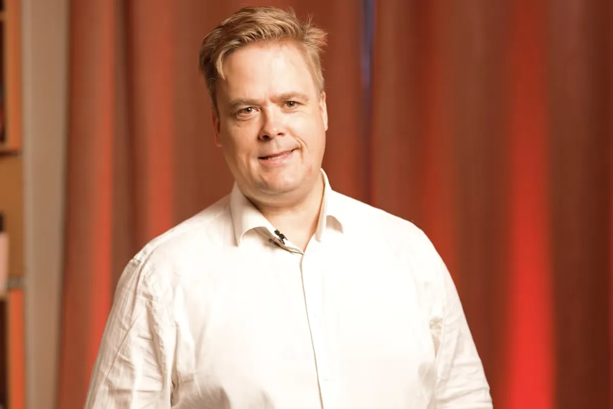 Man with light brown hair wearing a white shirt, smiling in front of a red curtain background.