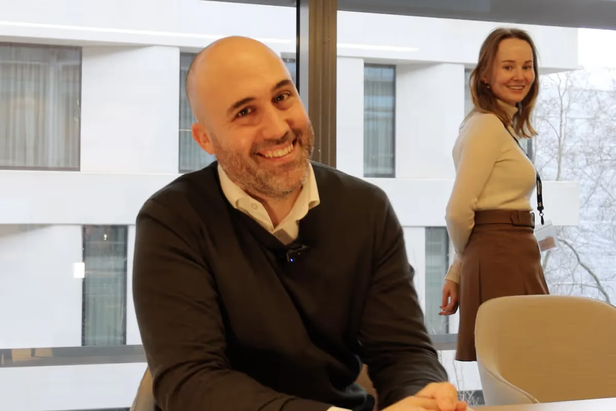 Smiling bald man in dark sweater sitting at a table with a smiling woman in a beige top and brown skirt standing behind him by a window.
