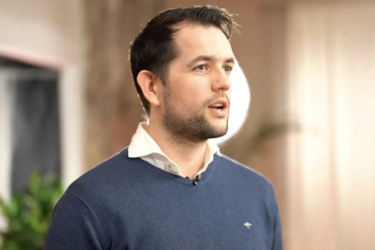 Man with short dark hair and beard wearing a white collared shirt and blue sweater, speaking indoors with blurred background.