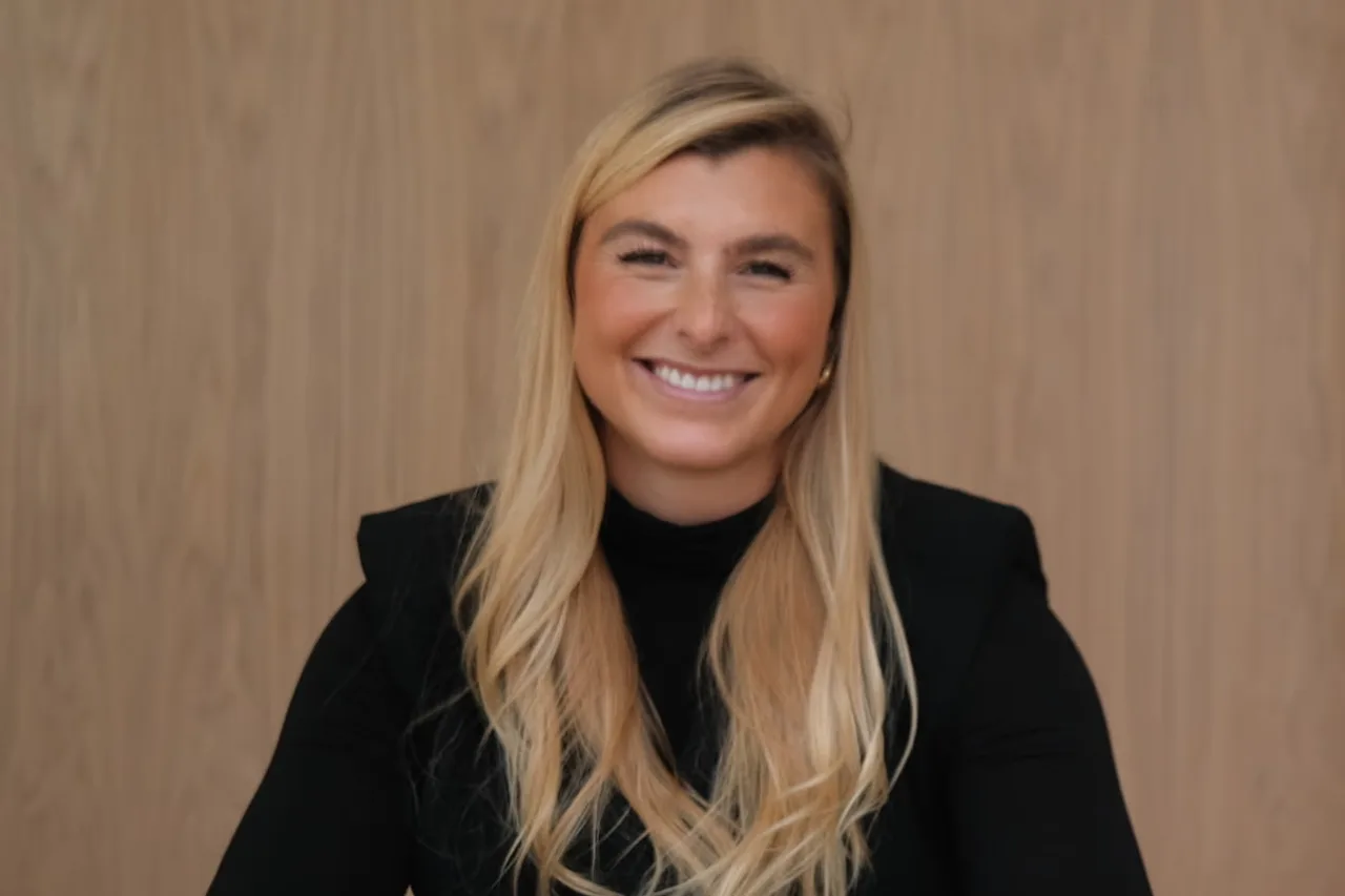 Smiling woman with long blonde hair wearing a black top against a wooden background.