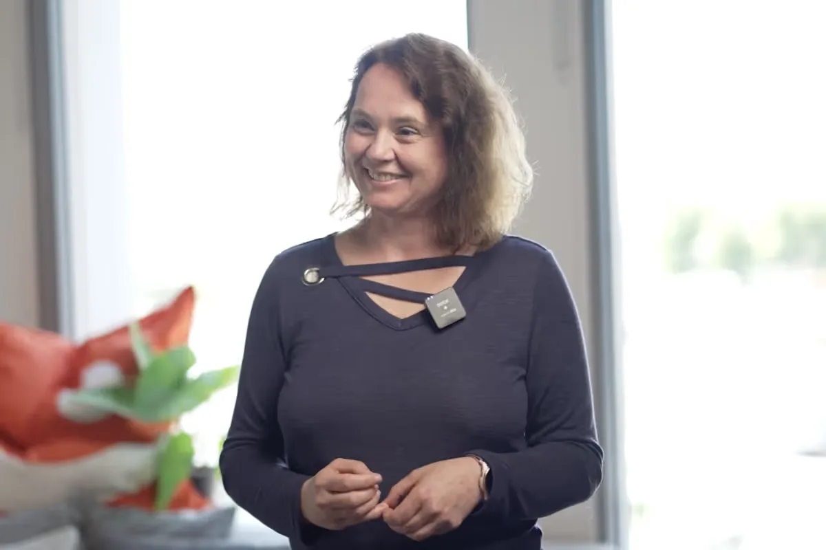 Smiling woman wearing a dark long-sleeve shirt and a small microphone clipped to her neckline, standing indoors with a blurred plant and cushion in the background.
