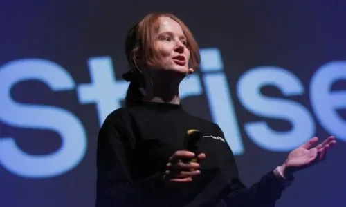 Woman with red hair speaking on stage holding a remote, with a blurred background text starting with ‘Strise’.