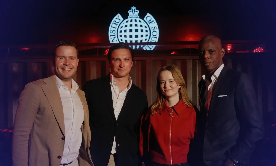 Four people standing together in a dimly lit setting with a Ministry of Sound logo illuminated in the background.